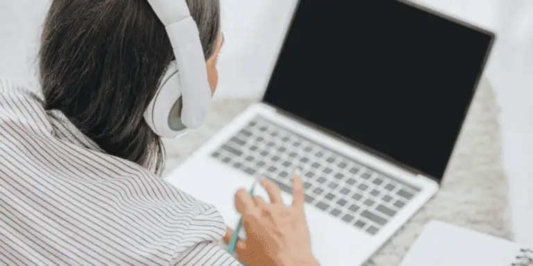 A person wearing white headphones uses a laptop with a blank screen, viewed from behind and slightly above. Their hand is on the trackpad as they sit on a light-colored surface, ready to begin virtual training.