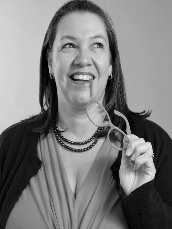 Elena Gonzalez smiles, looking upward while holding eyeglasses near her mouth. She has shoulder-length hair, wears a necklace, earrings, and a blouse with a cardigan. The photo is in black and white.