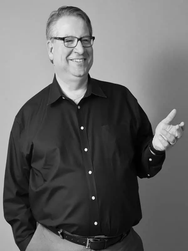 Bob Dianetti, a middle-aged man wearing glasses and a dark button-up shirt, smiles and gestures with one hand while standing against a plain background.