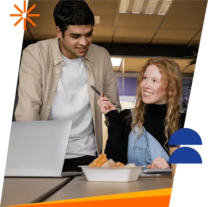 A woman with long red hair smiles and gestures with a pen while talking to a man standing beside her. They are at a table with a laptop and a container of food in a modern office space.